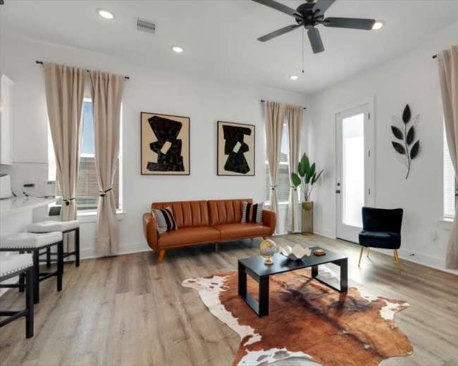 The room features a modern living space with a brown leather sofa, two black chairs, and a coffee table placed on a decorative rug, complemented by large windows with white curtains.