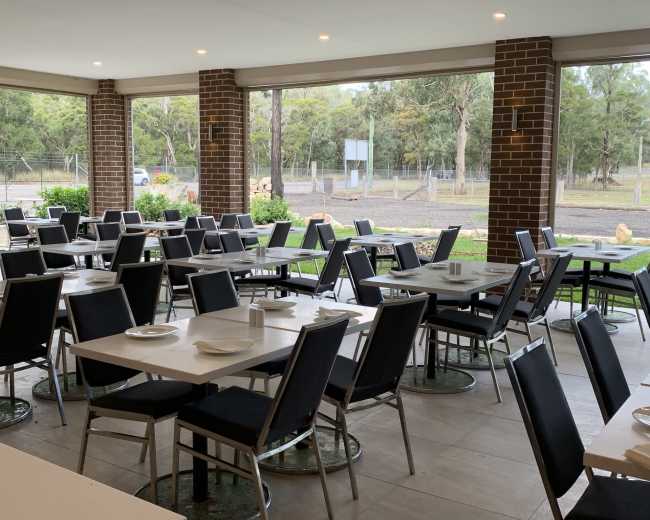 A spacious dining area with gray tables and chairs arranged neatly, surrounded by large windows overlooking a green landscape.