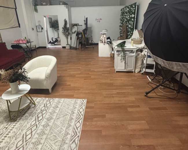 The image depicts an interior space with wooden floors, featuring a white armchair, a table, decorative plants, and a photography backdrop.