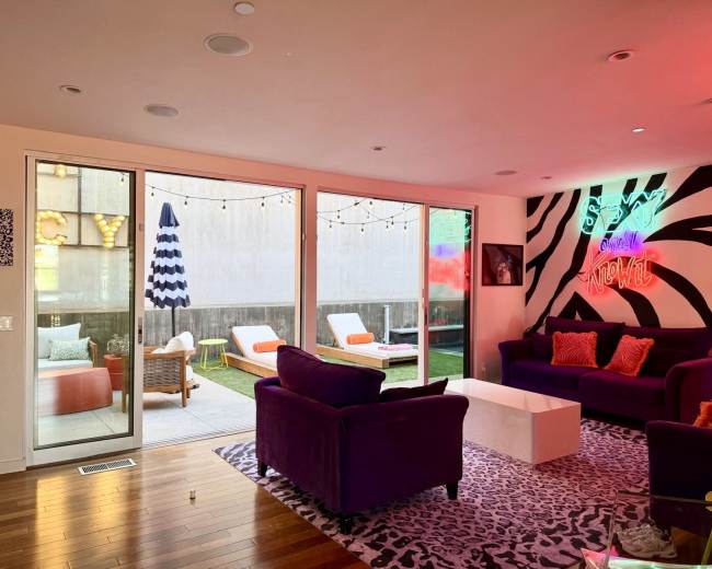 The room features a vibrant seating area with purple couches and a white coffee table, alongside large glass doors leading to an outdoor space with colorful lounge furniture and string lights.