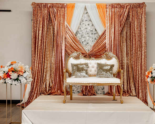 An elegant event setup featuring a white and gold seating area with plush cushions, flanked by vibrant floral arrangements and draped sequined backdrops in shades of rose gold and white.