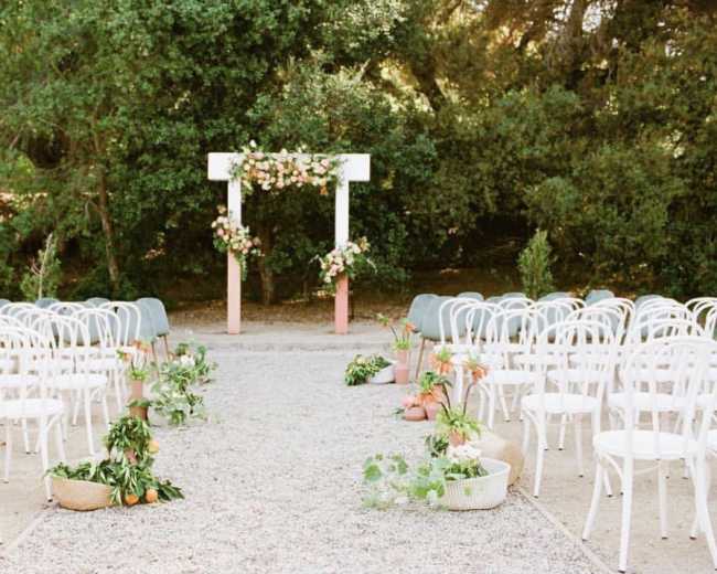 A wedding ceremony setup features white chairs arranged in rows facing a floral arch surrounded by greenery.