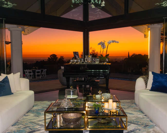 The image shows a modern living room with two white sofas facing a glass wall that offers a view of a sunset over a landscape.
