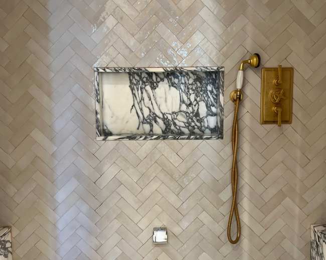 A modern shower with herringbone-patterned tiles, a marble shelf, and a gold shower fixture.
