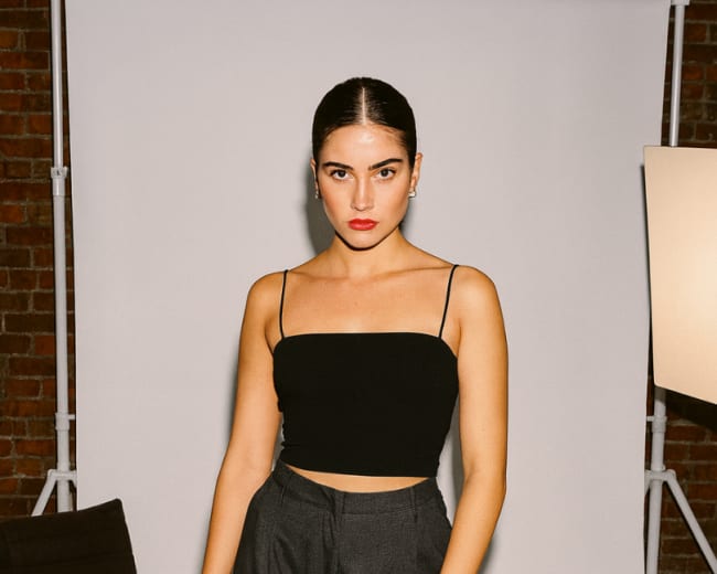 A woman stands confidently in a black crop top and gray trousers against a neutral backdrop in a studio setting.