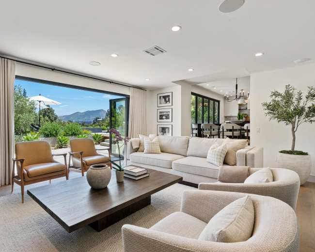 A modern living room with a light-colored sofa and chairs, a wooden coffee table, and large glass doors that open to a patio with a view of mountains and greenery.