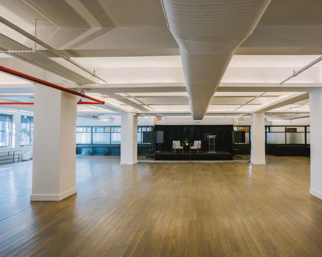 The image shows a spacious, empty office area with wooden flooring, large windows, and a small stage setup at the far end.