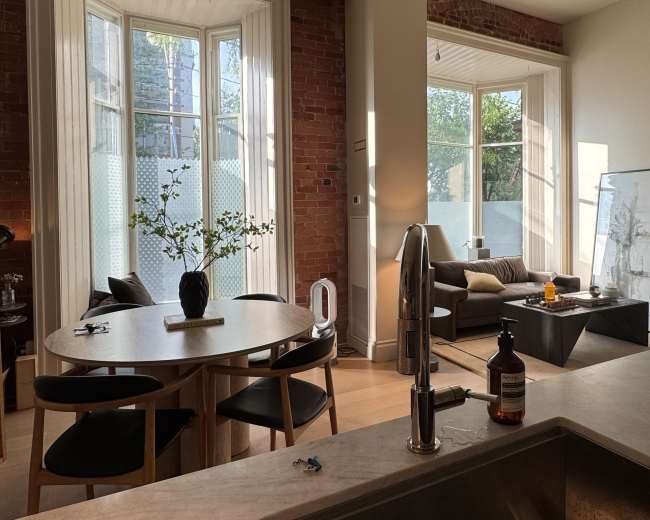 The image shows a modern living space with large windows, a dining area featuring a round table and chairs, and a comfortable seating area with a couch.