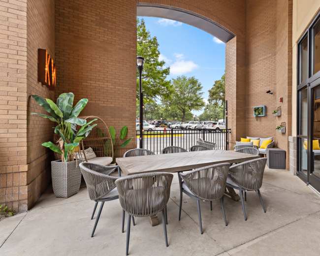 The image shows an outdoor dining area featuring a large table surrounded by grey chairs, with potted plants and a view of a parking lot beyond.