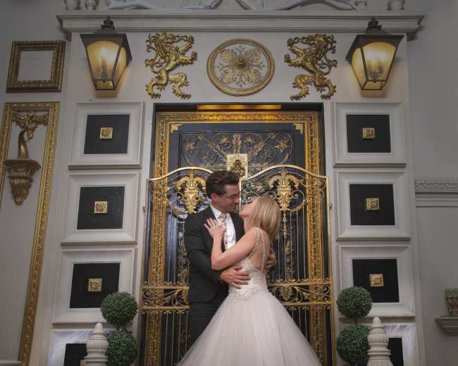 A couple wearing formal attire embraces in front of an ornate gold-and-black door adorned with intricate designs.