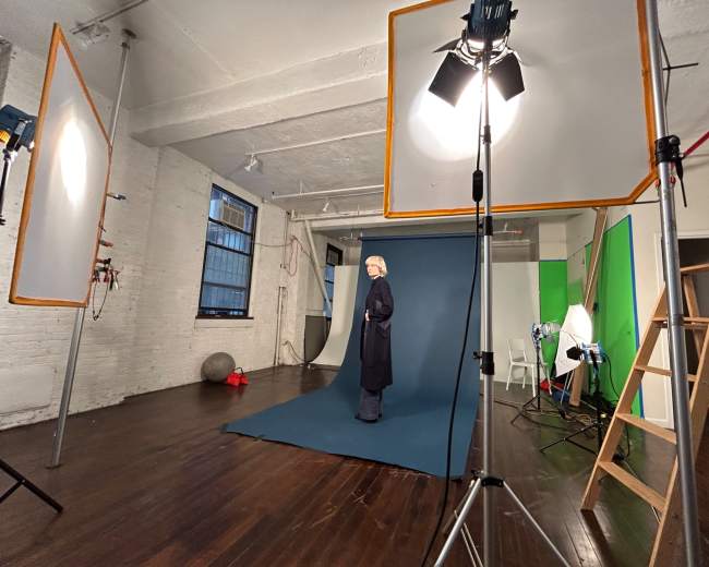 A model stands on a blue backdrop in a well-lit photography studio, surrounded by lighting equipment and props.