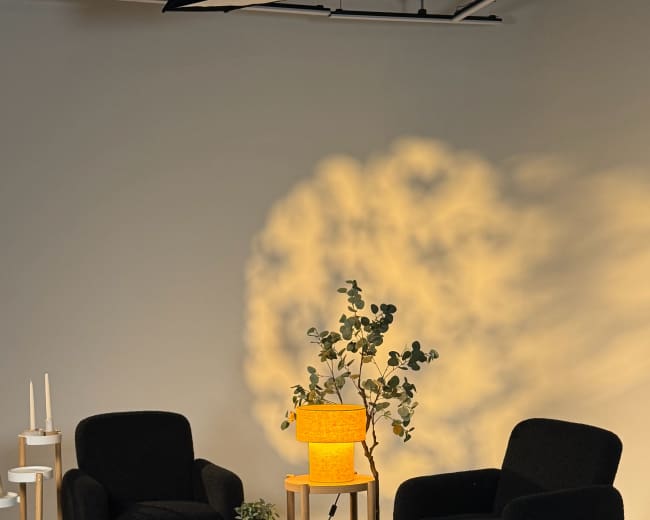 The image features two black armchairs on a textured rug, a round wooden coffee table, a yellow lamp, and a small table with candles beside a potted plant in a softly lit indoor setting.