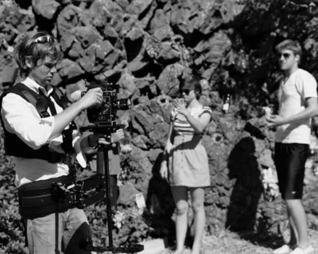 A filmmaker adjusts his camera equipment while two individuals stand nearby, one holding a drink and the other examining a piece of equipment.