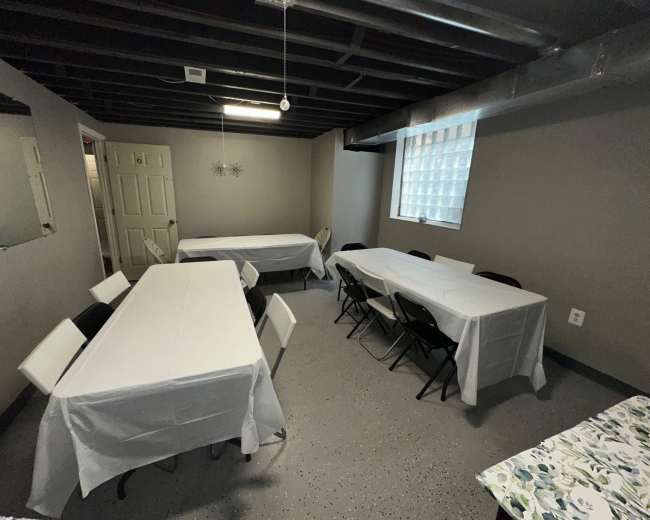 A basement room with several long tables covered in white tablecloths and chairs arranged around them, along with a mirror and a window featuring a glass block design.
