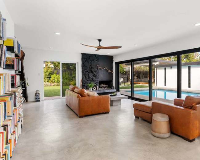 The modern living room features two leather sofas facing a media area, with large glass doors opening to a poolside view.