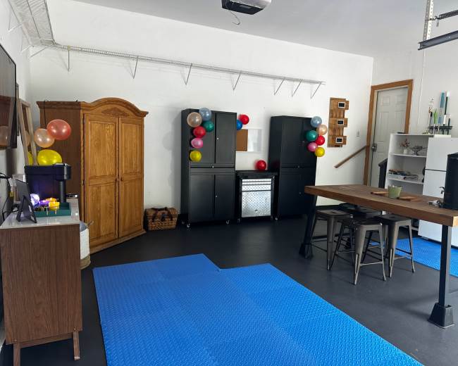 The image shows a garage converted into a party space, featuring a large wooden table, black cabinets with colorful balloons, and a blue rubber floor mat.