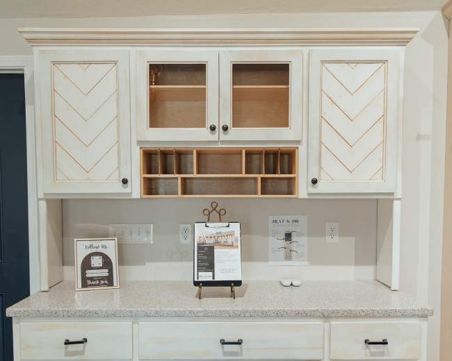 The image shows a modern kitchen cabinet unit with a light-colored wood finish, featuring chevron-patterned doors and glass-front sections, situated against a wall with a countertop and decorative items displayed.