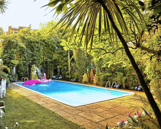 The image shows a private swimming pool surrounded by lush greenery and tropical plants, with pink floaties in the water and white chairs positioned nearby.