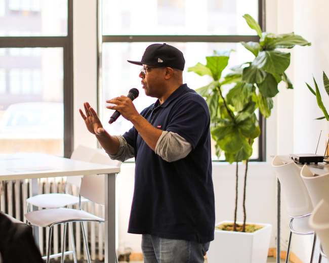 A man talking into a microphone stands in a bright room with large windows and plants.
