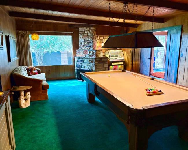 A cozy game room features a billiards table, a stone fireplace, and a sofa by a window.