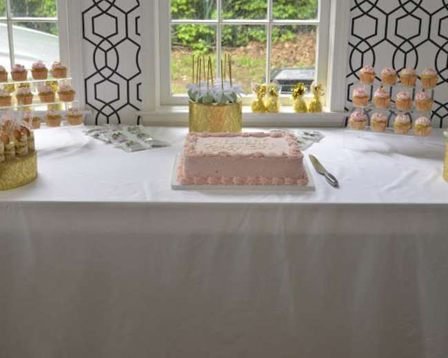 A decorated table features a pink frosted cake at the center, surrounded by trays of cupcakes and dessert treats.