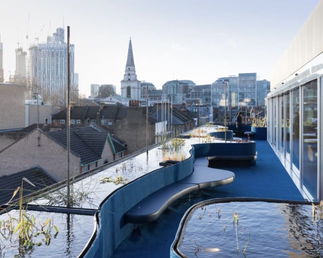 A rooftop terrace with wavy seating along a water feature, overlooking a city skyline with modern buildings and a church spire.