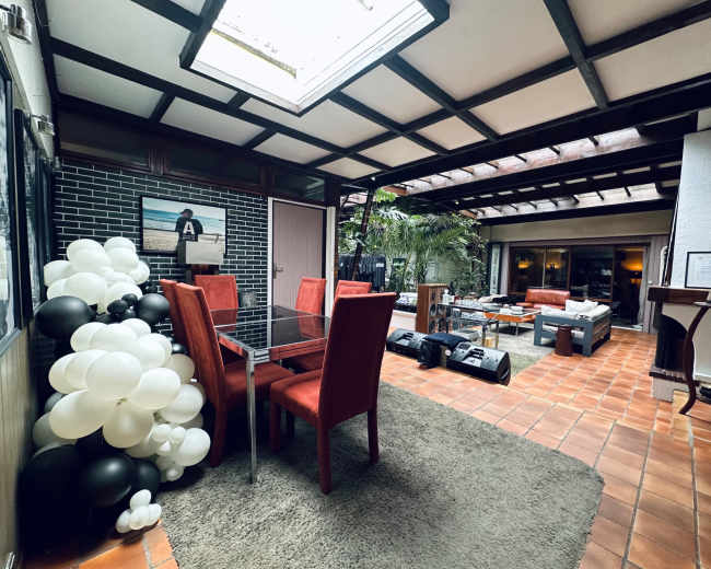 The image shows a well-lit indoor courtyard featuring a dining table with red chairs, a seating area with a couch, and decorative elements including black and white balloons.