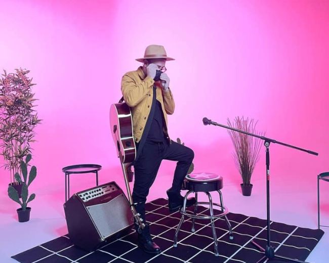 A musician sits on a stool with a guitar in a room accented by pink lighting, surrounded by potted plants and a microphone on a stand.