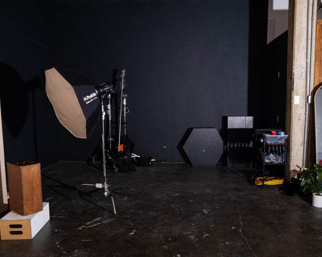 The image depicts a dimly lit photography studio featuring lighting equipment, a large mirror, and various props against a dark wall.