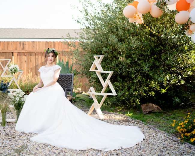 A woman in a white wedding dress sits on a chair in a garden decorated with geometric wooden sculptures and balloons.