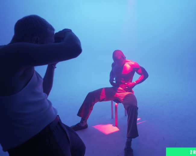 A photographer captures an artistic shot of a male model seated on a stool under colorful lighting in a foggy environment.