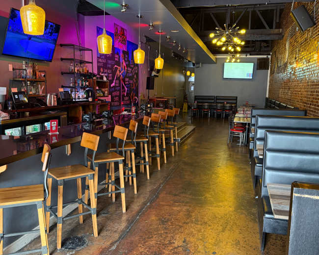 The image shows the interior of a modern bar with a long counter, several empty chairs, a brick wall, and multiple screens displaying images.