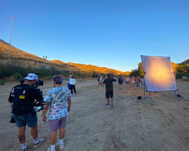 A film crew sets up a shoot in a mountainous area at sunset, with several models positioned in front of a large reflector.