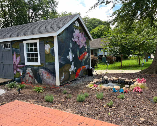 A shed features a mural of koi fish and water lilies, surrounded by a landscaped garden with flowers and stones.