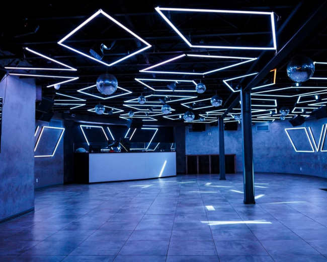 The image shows an empty nightclub with a geometric ceiling design featuring blue LED lights and mirrored disco balls.
