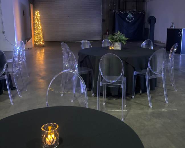 A spacious indoor venue features round tables with black tablecloths and transparent chairs, along with a small illuminated tree in the corner.