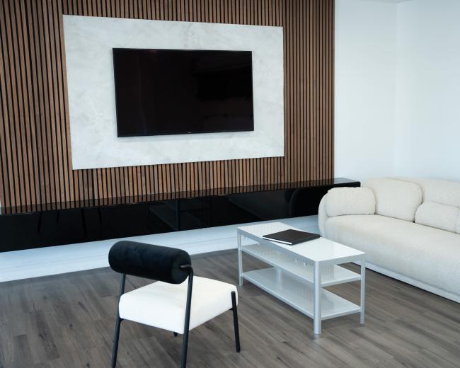 The image depicts a modern living room with a plush sofa, a small coffee table, and a wall-mounted television framed by a textured panel.