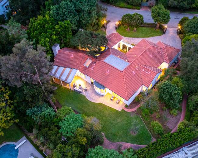 An aerial view of a single-story house with a red tile roof surrounded by lush greenery and a circular driveway.