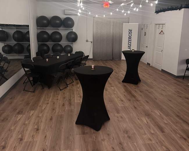 A spacious room with wooden floors, black folding chairs arranged around two tall tables, and decorative string lights overhead, along with a wall featuring large black exercise balls.