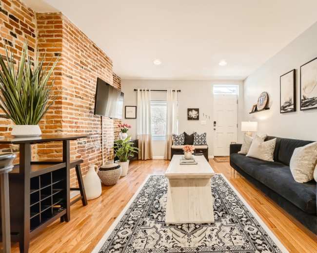 The image shows a modern living room featuring a black sofa, a wooden coffee table, a textured brick wall, and decorative artwork.