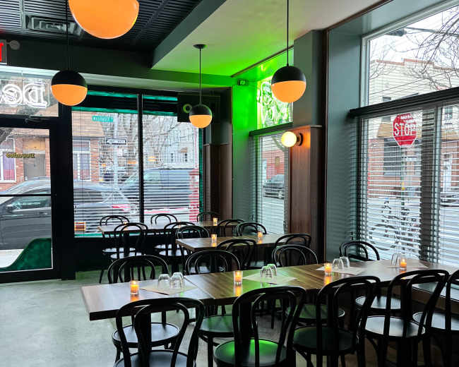 The image shows a modern restaurant interior featuring rows of wooden tables and black chairs, illuminated by round pendant lights.