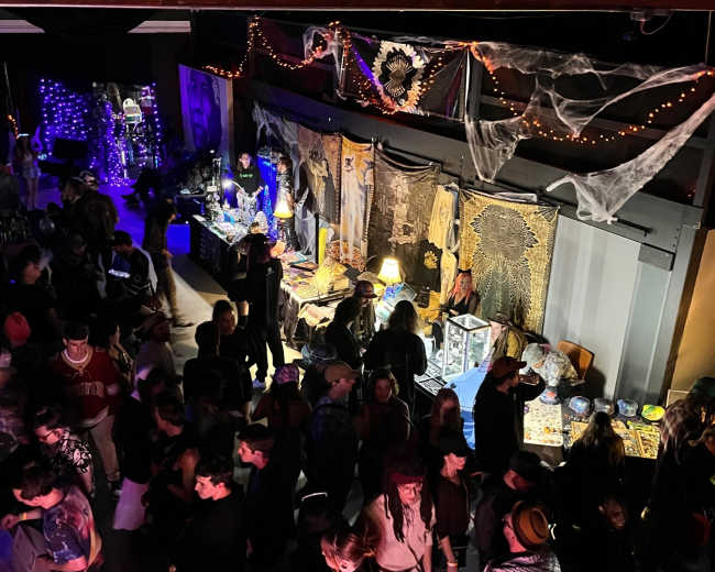 A crowd of people gathers in a dimly lit party venue decorated with spider webs and various props.