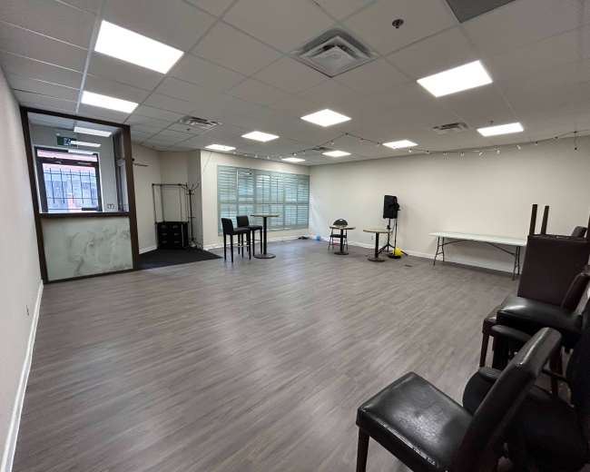 The image shows a spacious, empty room with wooden flooring, a few tables and chairs, and large windows allowing natural light to enter.