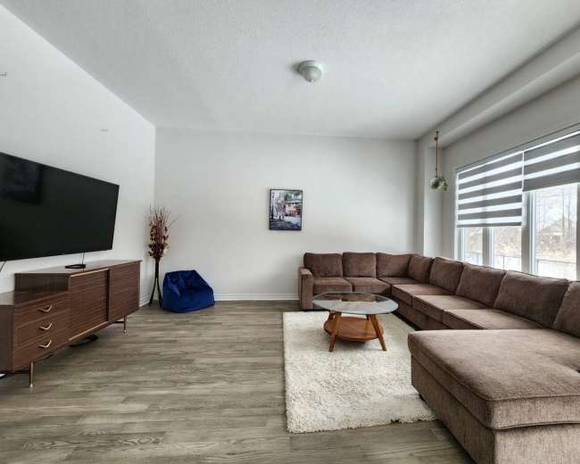 A spacious living room features a large sectional sofa, a round coffee table on a rug, a television mounted on the wall, and ample natural light from the windows.