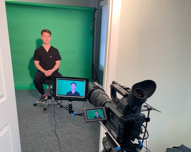 A person sits on a stool in front of a green screen while being filmed by a camera on a tripod.