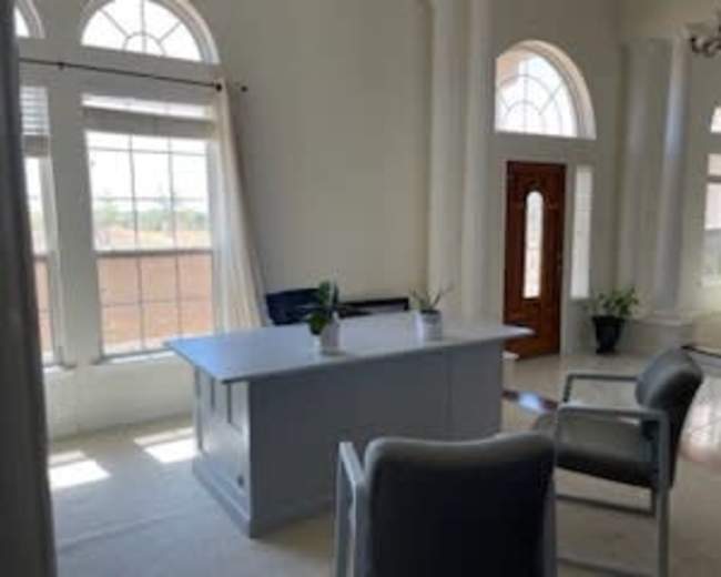 The image shows a well-lit office space featuring a large desk, two chairs, and large windows with white curtains.