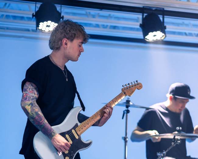 A guitarist performs on stage with a drummer in the background during a live music event.