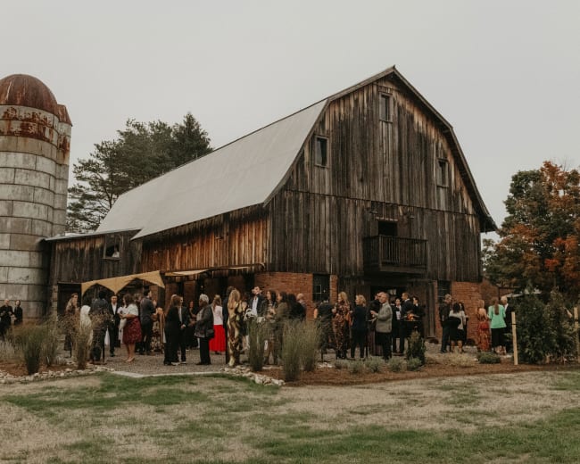 A gathering of people is taking place outside a rustic barn, with a silo nearby and trees in the background.