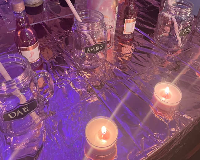 The image shows a kitchen countertop decorated with pink champagne, jars filled with colorful liquids, cupcakes, and lit candles.