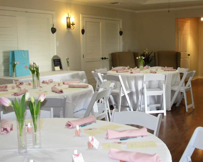 A well-lit event space set up for a gathering, featuring white tablecloths on round tables with pink napkins, vases of tulips, and a few chairs arranged around the space.